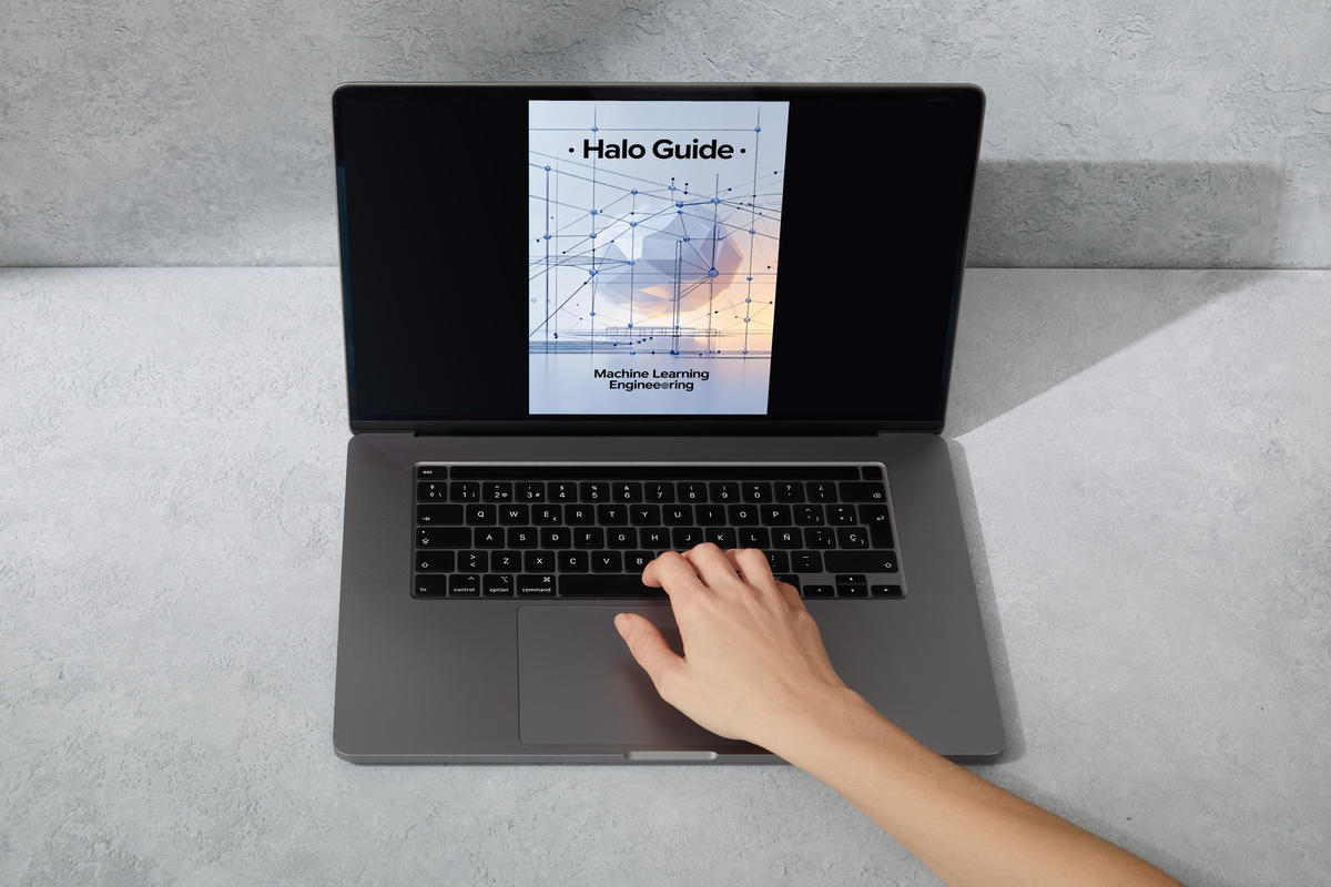 A gray laptop sits on a light gray surface with a hand resting on the trackpad. The screen displays a book cover titled 'Halo Guide: Machine Learning Engineering'.