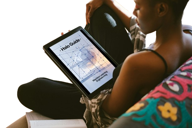 A person sitting indoors holds a tablet displaying a book or guide titled "Halo Guide: Modern Learning Engineering." The scene is brightly lit from the left, with the person wearing black leggings and a plaid top, and a colorful pillow visible in the foreground.